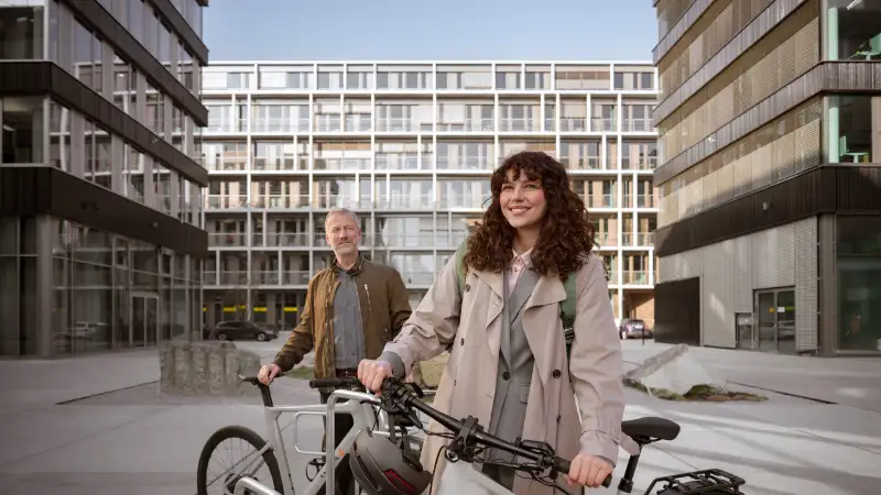 JobRadlerin steht mit Fahrrad abfahrbereit auf einem Firmencampus, im Hintergrund Kollege mit Fahrrad